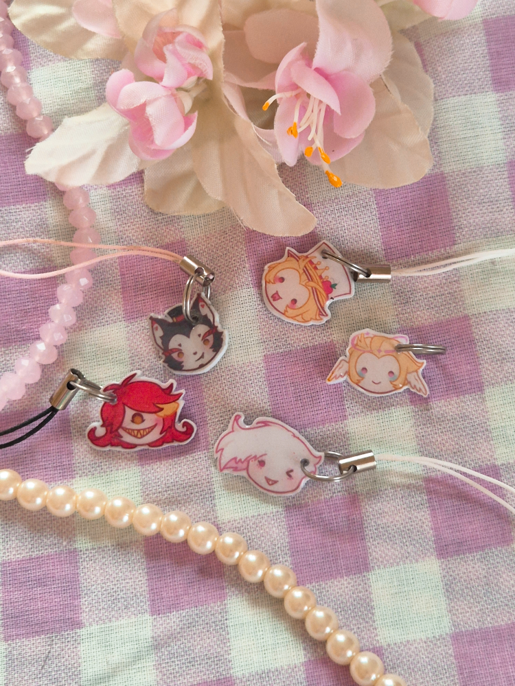 phone charms of Niffty, Husk, Lucifer, Angel and pre-fall Lucifer from Hazbin Hotel, fake flowers above them, strings of pearls next to them.