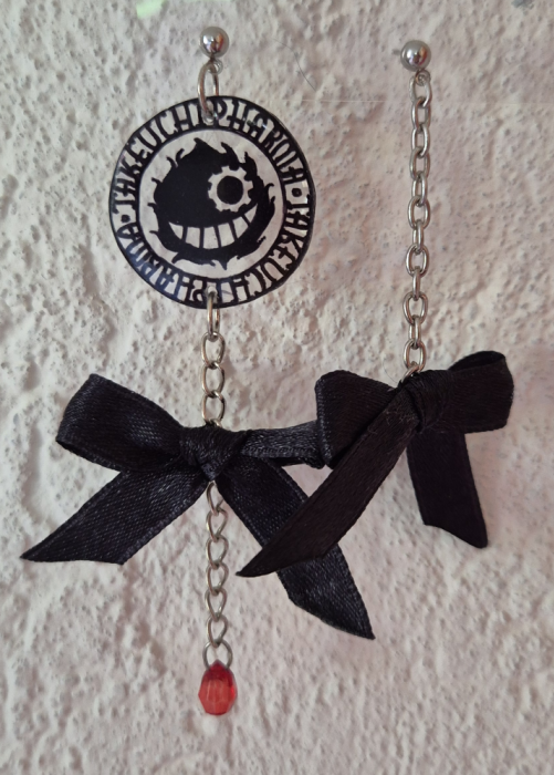 A pair of earrings suspended on clear plastic in front of a wall. The earrings are of a black logo with a grinning character that wears an eyepatch. Text around the head reads 'takeuchi pharma'. A bow dangled from it from a chain, a drop-shaped pearl at the bottom. The other earring is just a bow on a chain.